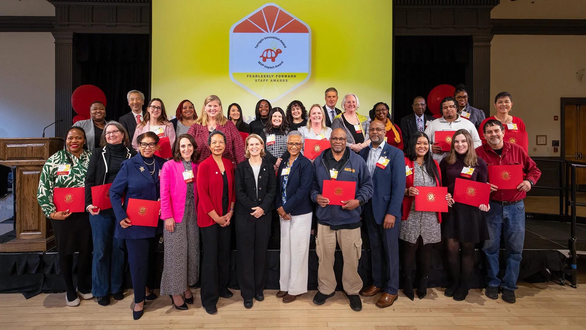 group of 25 award winner recipients, holding their certificate in front of a yellow screen with a logo, standing with university leadership